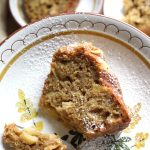 piece of apple cake sitting on china plate sprinkled with powdered sugar. Two small plates behind on the counter