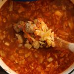A blue Dutch oven filled with the best sausage and sauerkraut soup with a wooden spoon scooping up the soup on a tea towel on counter