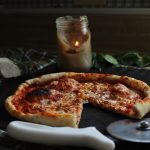 Sourdough pizza made with homemade sourdough pizza dough sitting on a black cast iron pizza stone with a tea towel underneath on counter. Pizza sauce in glass jar, mozzarella fresh herbs and a candle