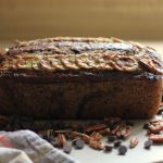 Sourdough Banana Bread sitting on a cutting board with pecans and chocolate chips and a tea towel on the countertop.