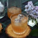 lilac lemonade in a glasses with lilac ice cubes sitting on a wooden coaster on a table cloth with fresh lilac flowers and a bottle of lilac lemonade in the background.