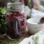 pickled beets prepared by getting sliced beets in a quart jar, crock of salt with salt spoon, beet leaves and rosemary sitting next to a tea towel on the counter top.